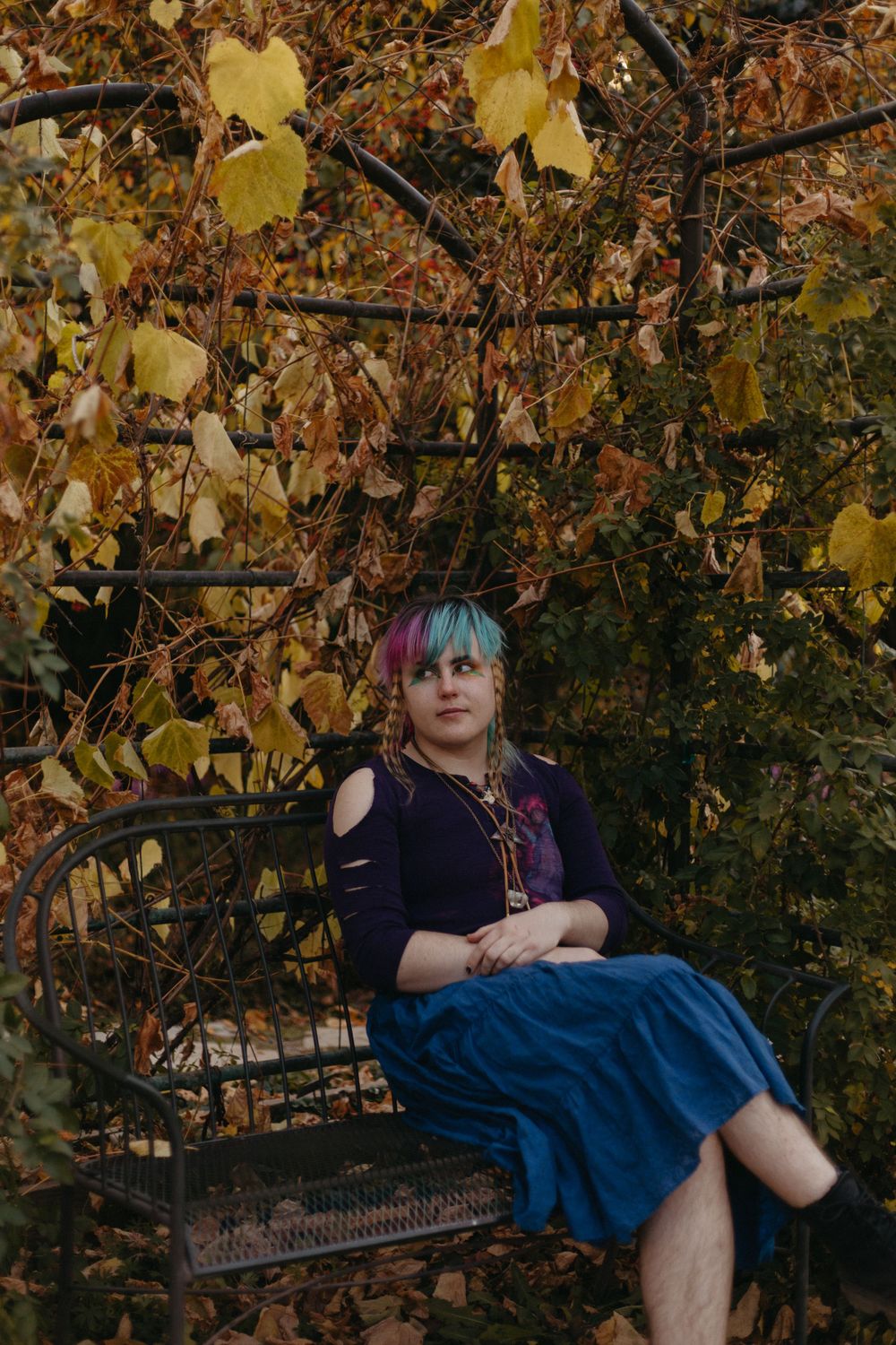 Person in a dark dress lounges against autumn foliage with golden leaves in a moody outdoor setting.