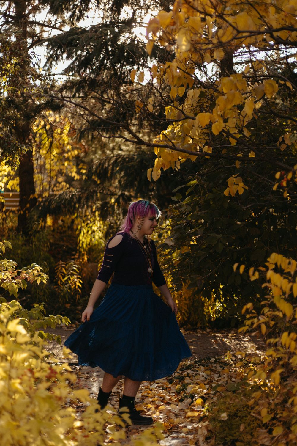 Figure in a dark dress crouches among fallen autumn leaves in a golden-lit forest scene.
