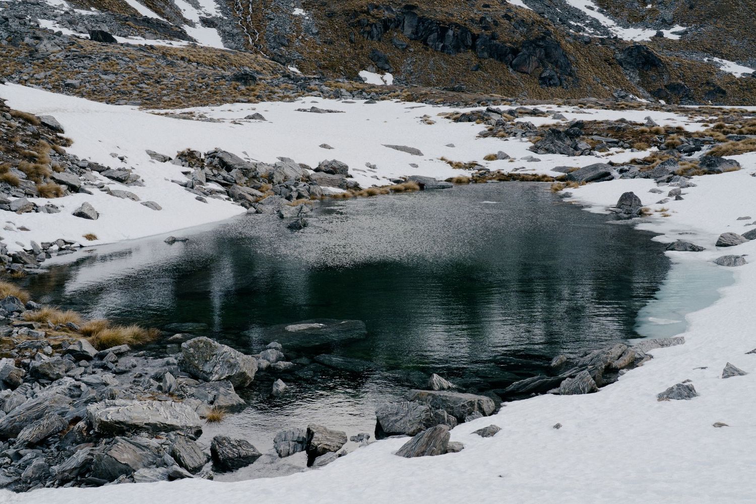 Snow-covered alpine lake surrounded by rocky terrain and patches of melting snow.