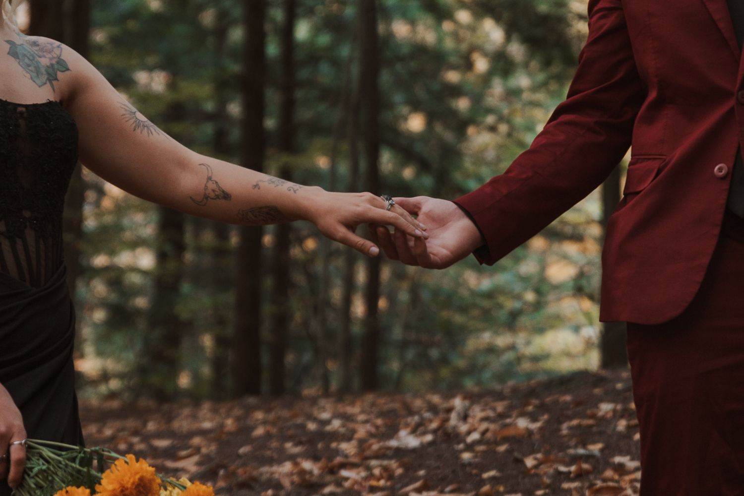 Two people hold hands in a romantic autumn forest setting with fallen leaves and burgundy clothing.