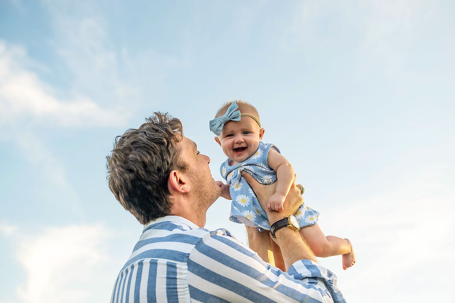 A parent lifts a happy child into the air against a bright blue sky background with clouds.