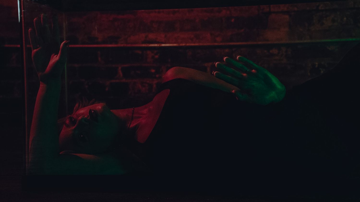 Person lying down in dramatic red and green neon lighting against a dark brick wall background.