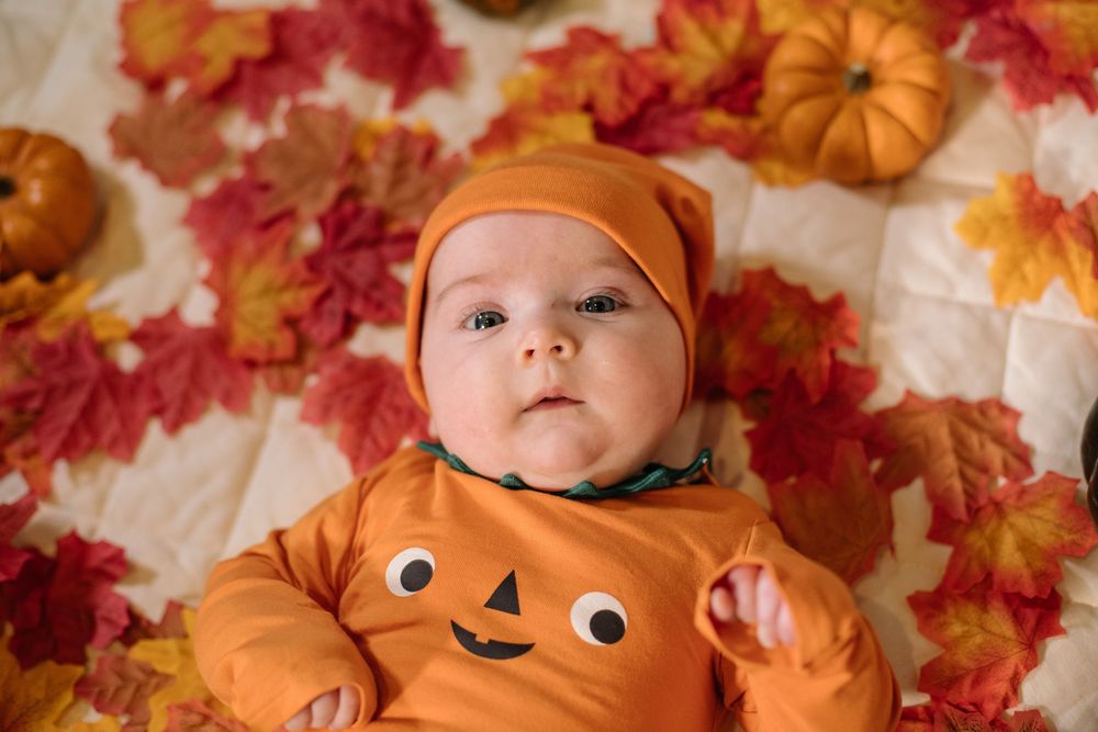 A baby wearing an orange pumpkin costume lies on a blanket surrounded by autumn leaves and small decorative pumpkins.