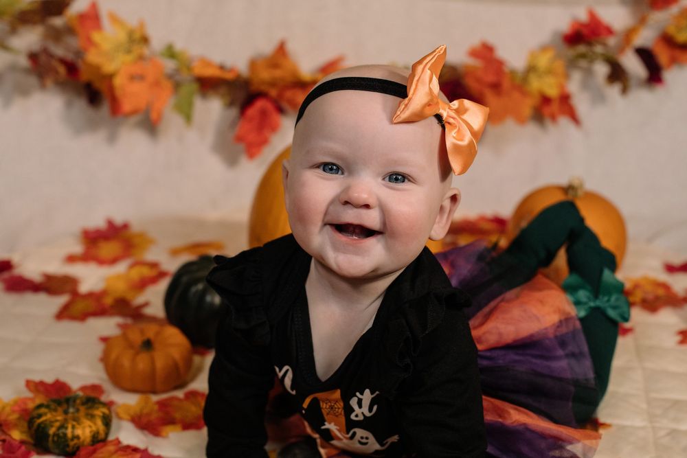 A smiling baby in a black outfit with orange bow sits among fall decorations with pumpkins and autumn leaves.