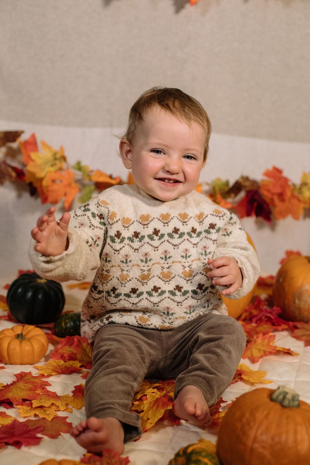 A smiling baby wearing a patterned sweater sits among autumn leaves and pumpkins in a festive fall photo setup.