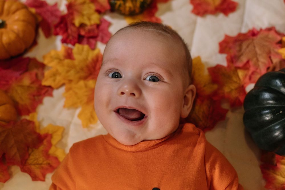 A smiling baby lies on a blanket surrounded by colorful autumn leaves and decorative pumpkins.