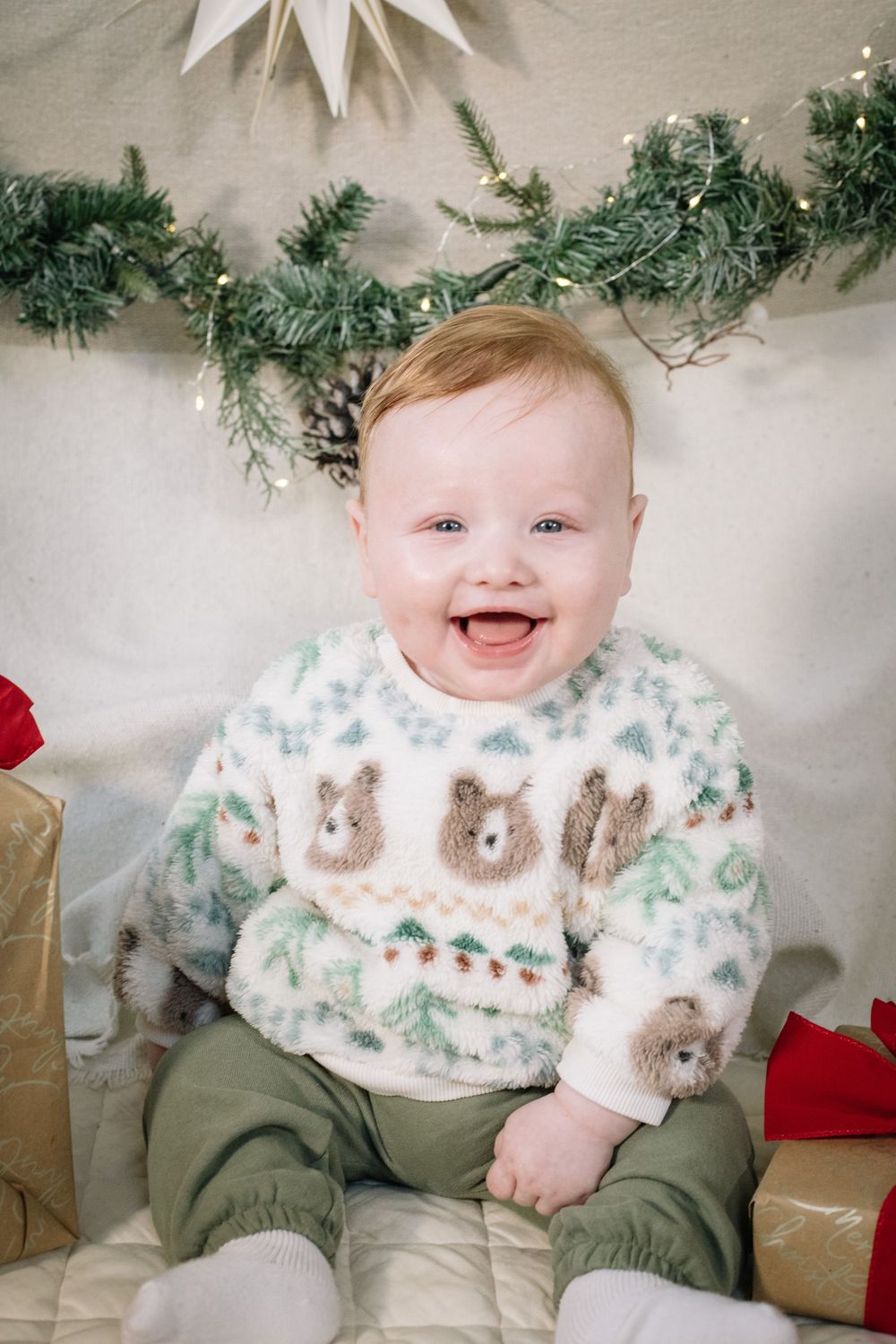 A baby wearing a festive white sweater with bunny designs sits in front of Christmas decorations and garland.