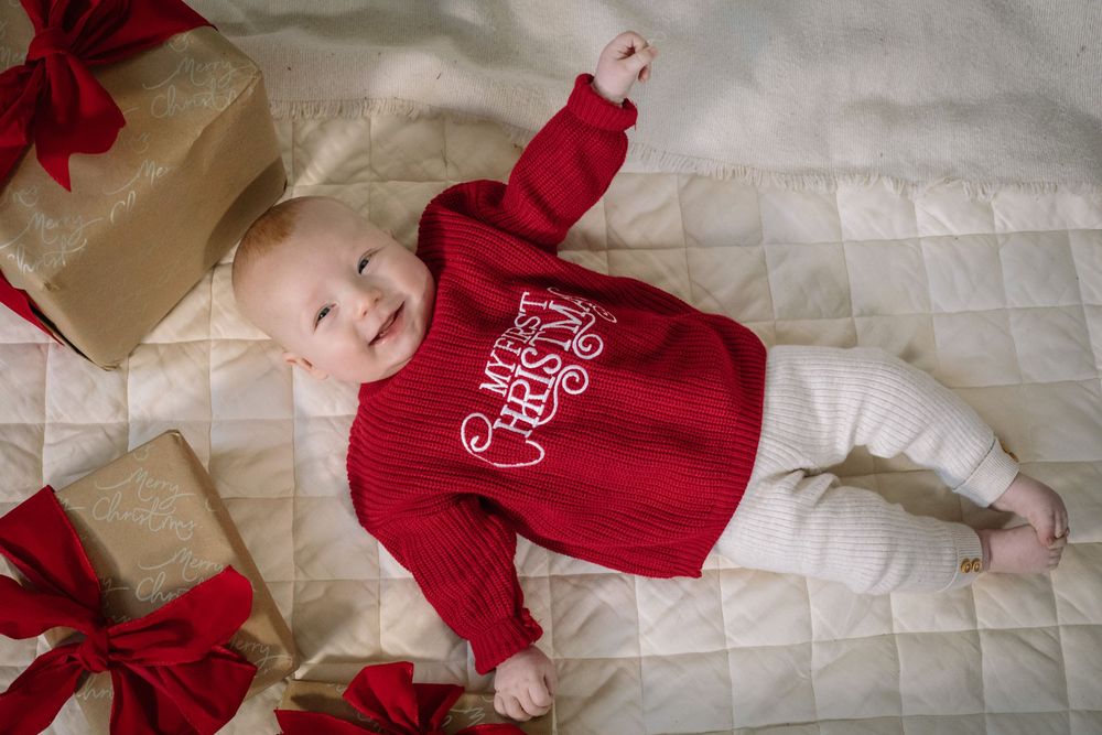A baby wearing a festive red holiday sweater lies on a quilted white surface next to wrapped presents with red bows.
