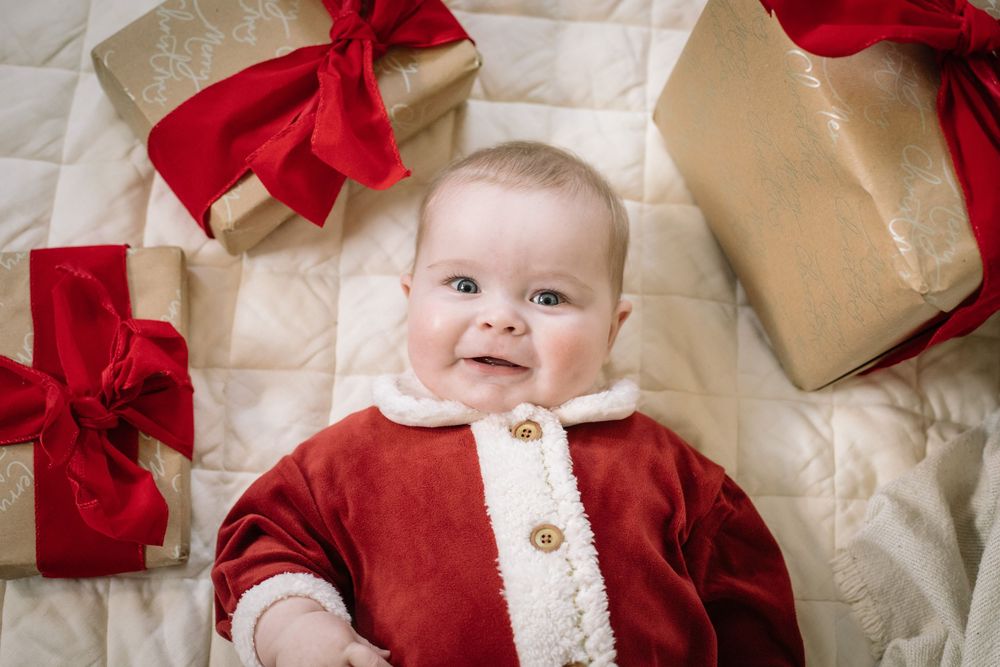 A baby in white and red holiday attire sits among wrapped Christmas presents with red bows.
