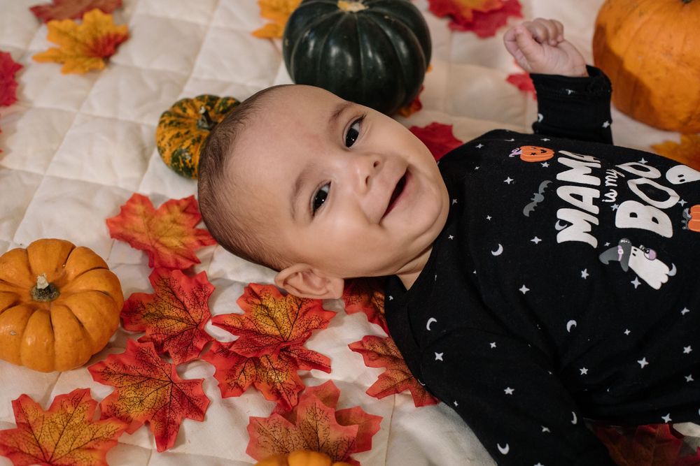 A smiling baby lies on white fabric surrounded by orange autumn leaves and pumpkins in a seasonal photo setup.