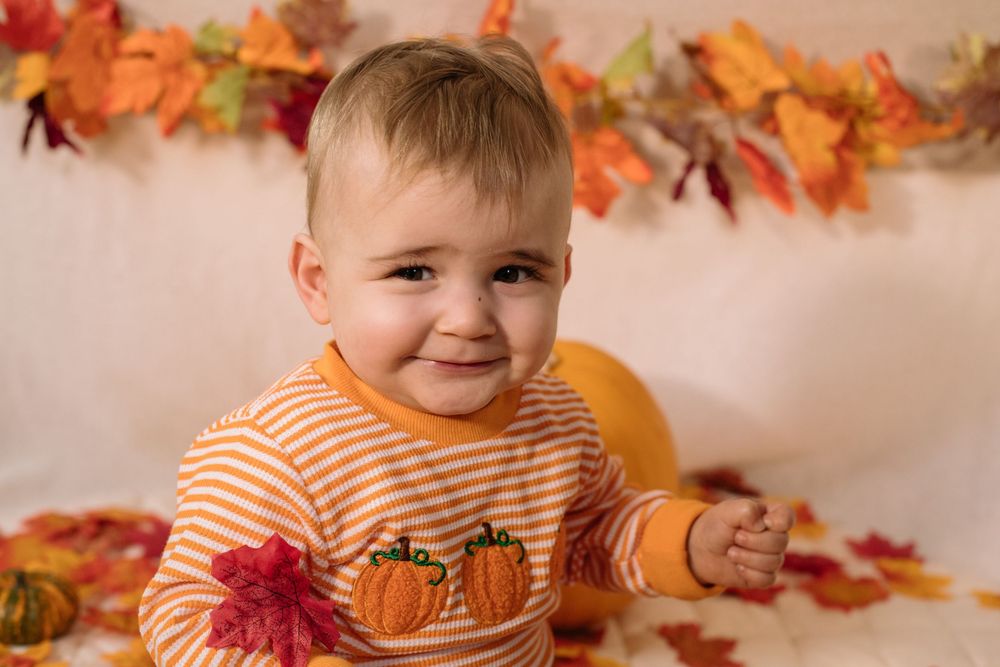 A young child in an orange striped pumpkin shirt sits with autumn leaves and decorations in the background.