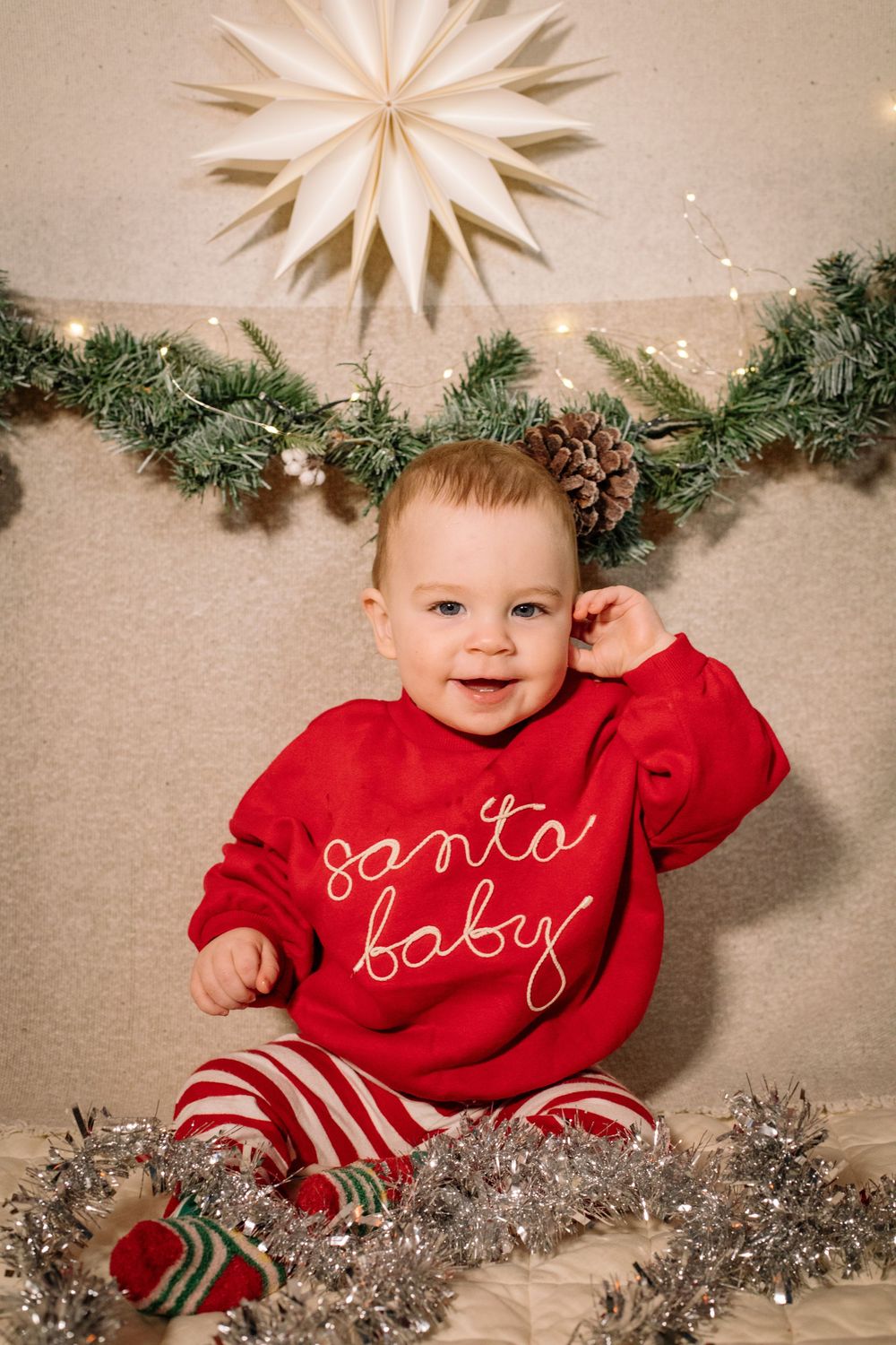 A baby wearing a red Santa-themed outfit lies under Christmas garland and decorative stars.