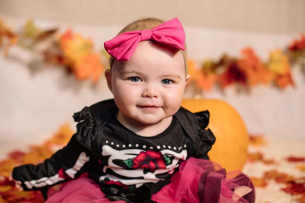 A child wearing a black dress and pink bow sits among colorful autumn leaves during a fall photo session.