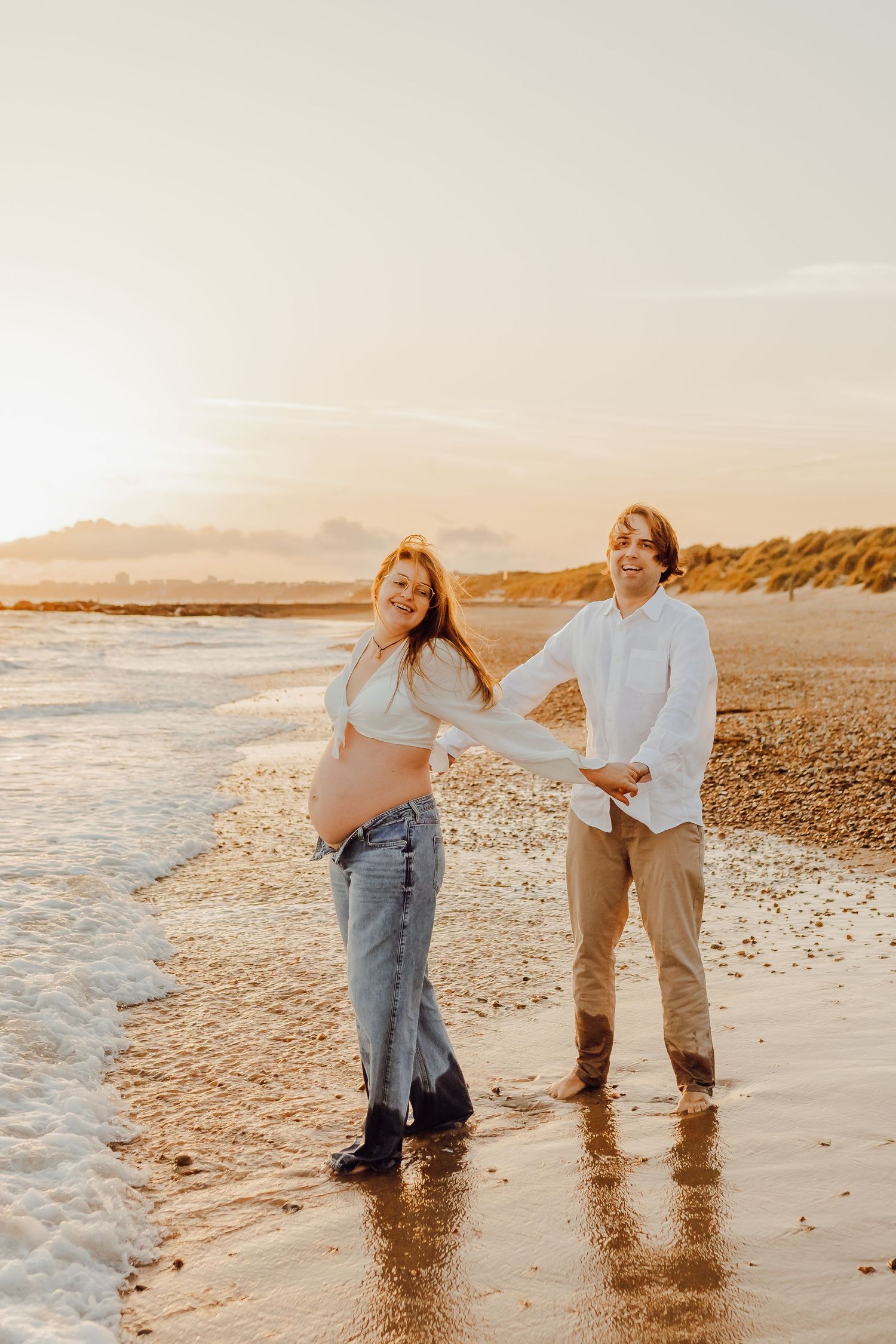 A pregnant couple strolling along the shoreline at Bournemouth, holding hands and laughing.
