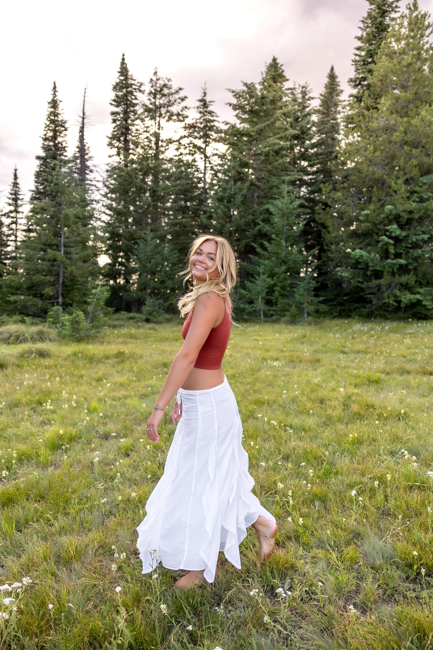 A person in a flowing white dress twirls through a mountain meadow filled with wildflowers and evergreen trees.