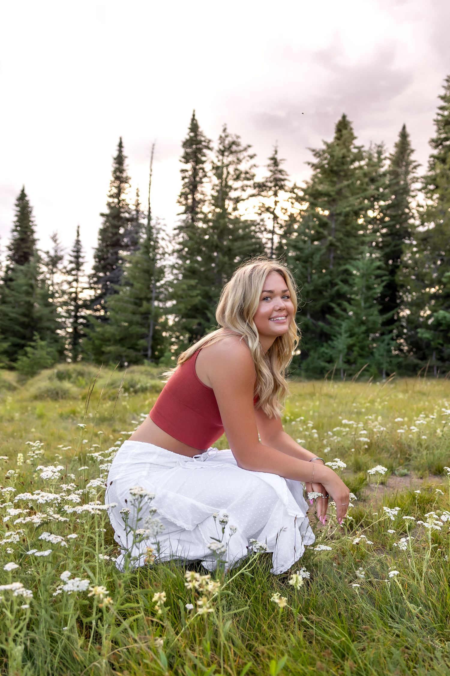 A person in a rust-colored top and white skirt poses joyfully in a wildflower meadow with evergreen trees in the background.