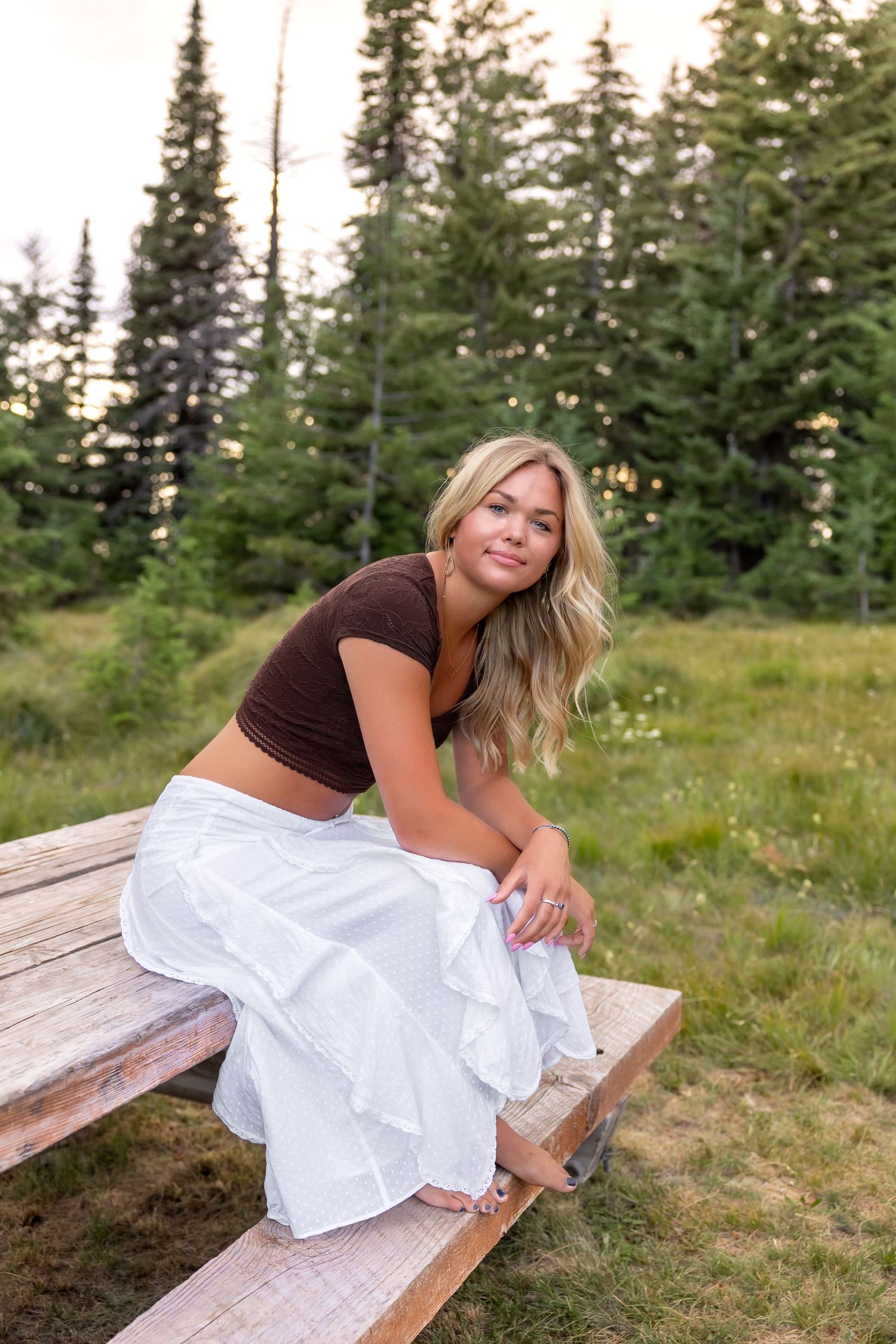 Someone in a white dress poses playfully on a wooden bench surrounded by evergreen trees and grass in nature.