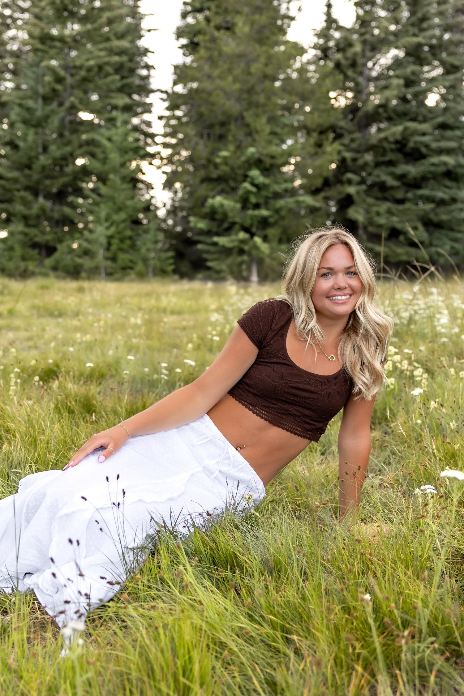 A person in a brown crop top and white skirt relaxes in a wildflower meadow with evergreen trees in the background.