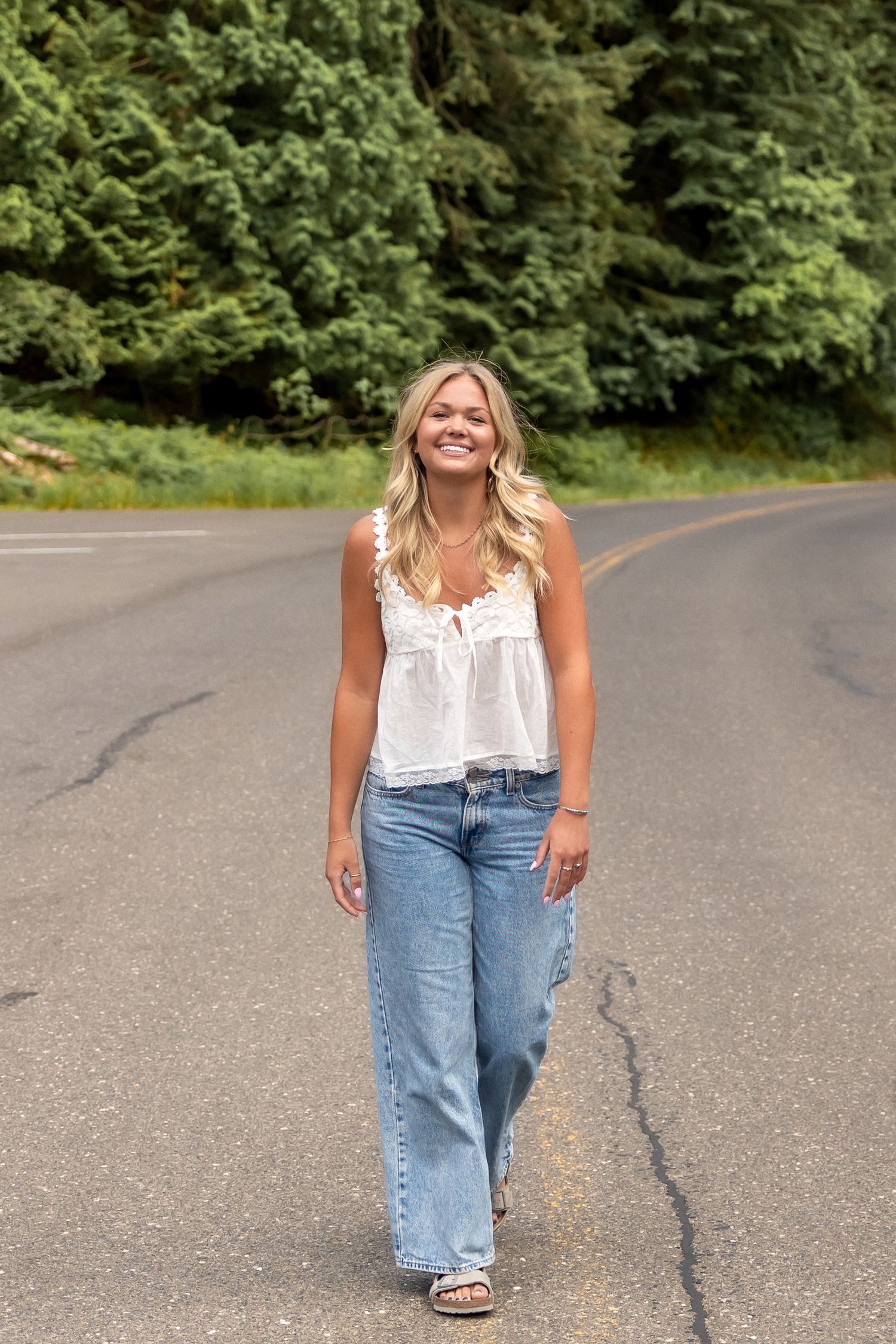 A casual summer outfit featuring a white sleeveless top and light blue jeans photographed on a paved road with trees.