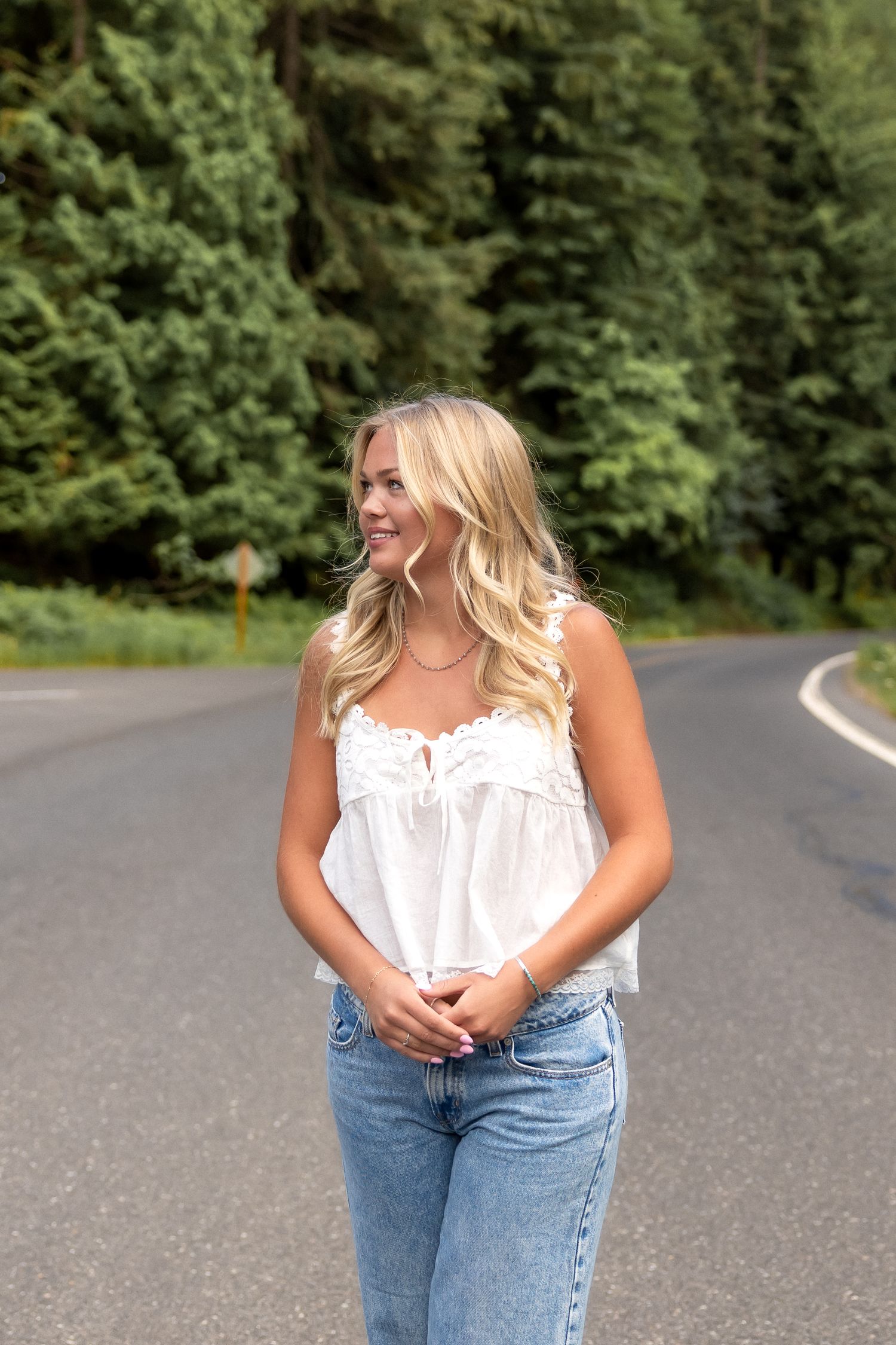 A woman in a white sleeveless top and jeans poses on a winding road surrounded by green trees.