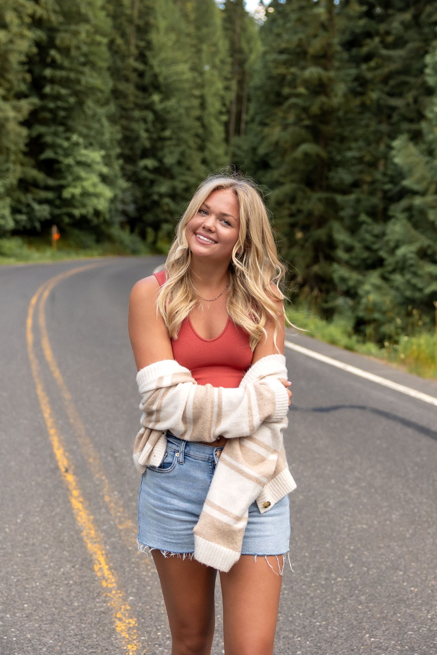 Woman in red tank top and denim shorts poses playfully on forest road wearing cream colored cardigan.