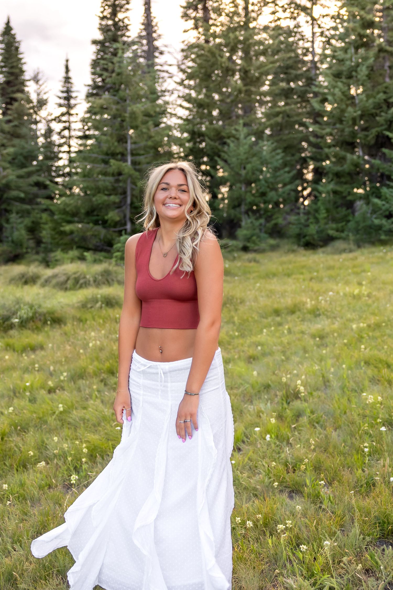 A person in a red top and flowing white skirt poses playfully in a mountain meadow with evergreen trees in the background.