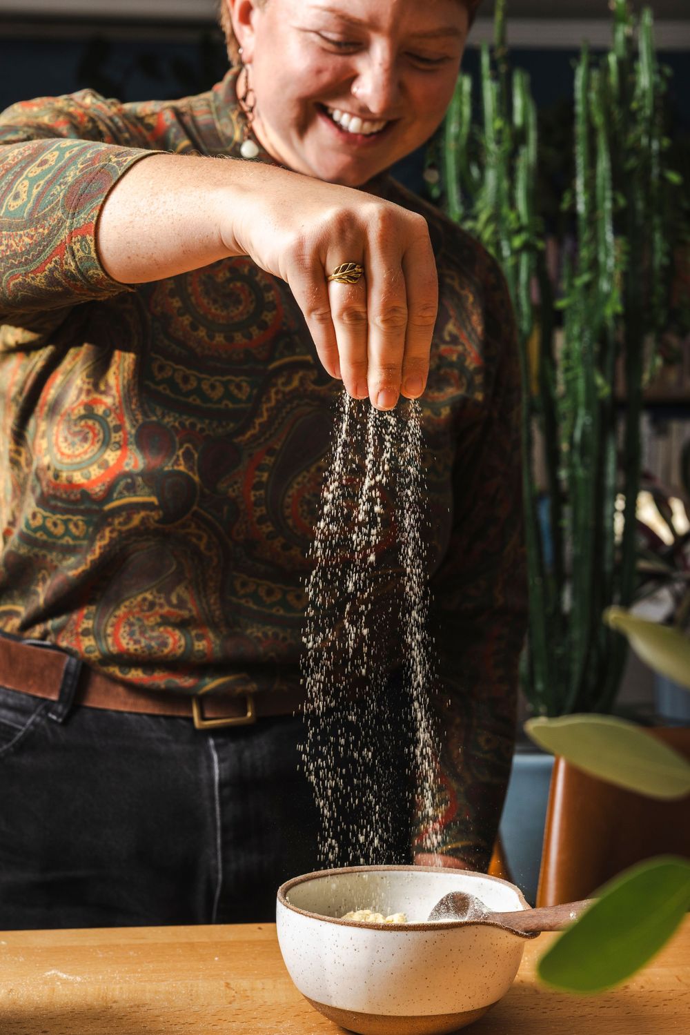 A person sprinkles seasoning into a white bowl while wearing a patterned shirt near houseplants.