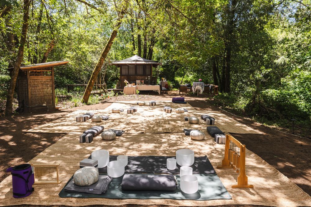 Outdoor meditation space with cushions and mats arranged on a wooden deck surrounded by trees.