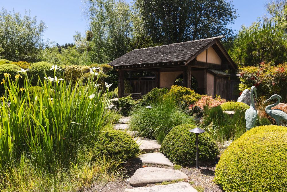 Winding stone pathway leads through a lush Japanese garden with ornamental grasses and traditional wooden structures.
