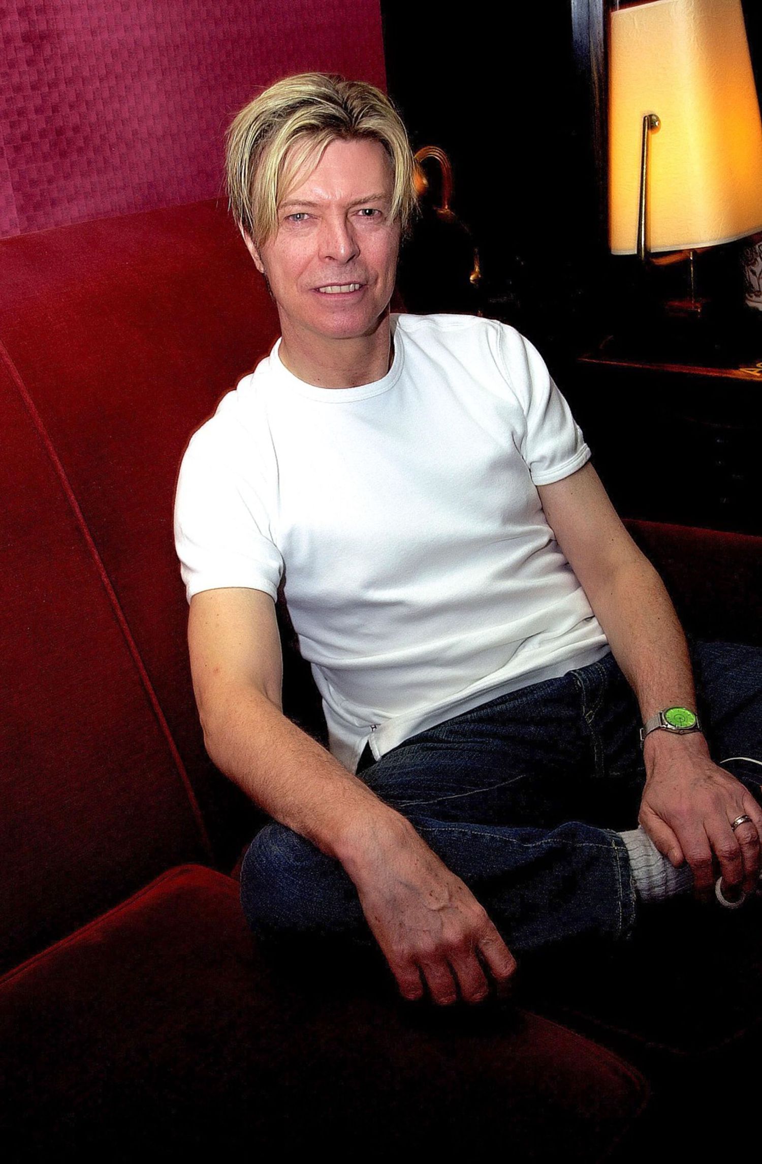 Person wearing a white t-shirt sitting on a dark couch in dim lighting with a lamp visible in background.