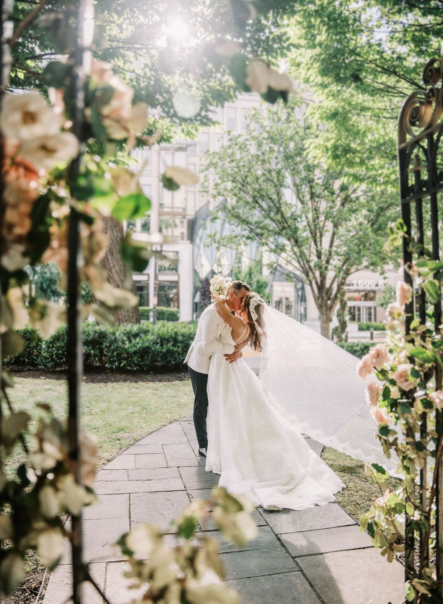 A bride in a flowing white dress twirls gracefully in a sunlit garden path surrounded by blooming trees and flowers.