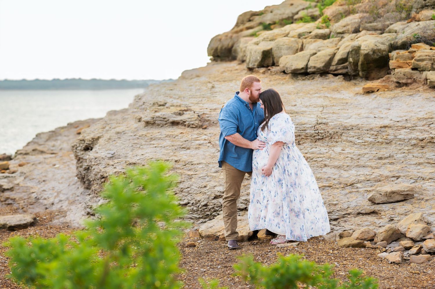 Soon-to-be parents at Rockledge Park next to the lake and large rock formation; dad kissing mom's forehead and with his hand on her bump