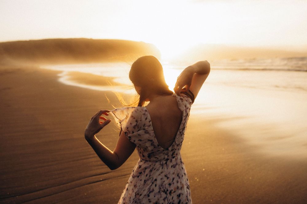 A person in a floral dress stands on a beach during golden hour sunset, silhouetted against the glowing sky.