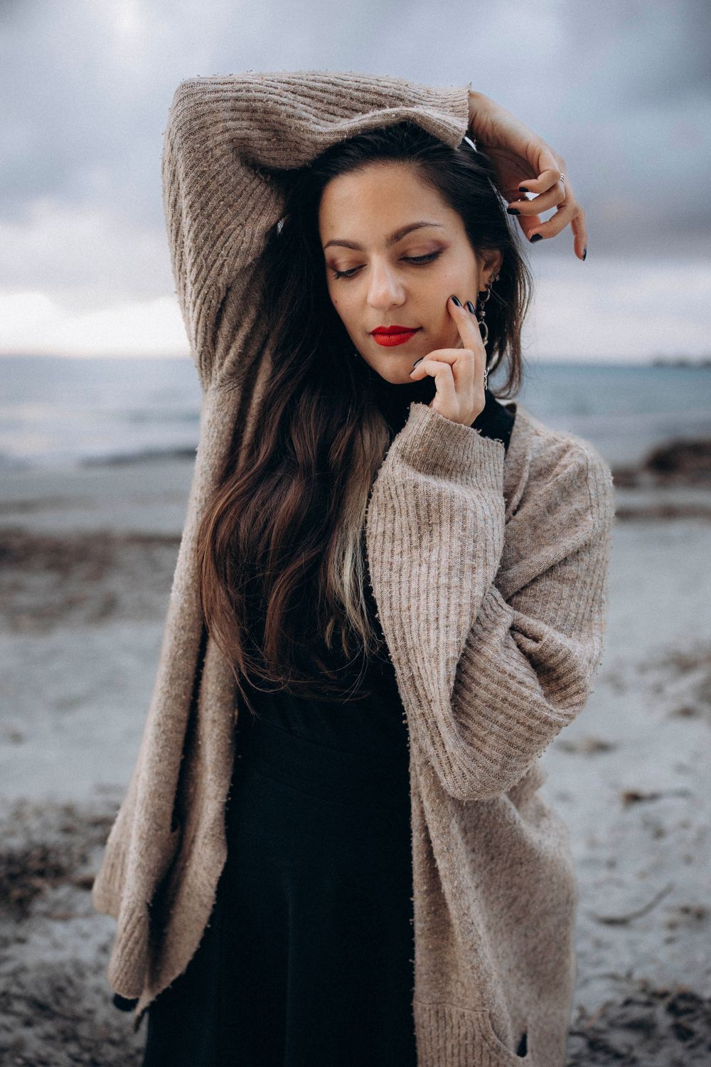 A person in a cozy beige sweater poses thoughtfully against a moody coastal backdrop on an overcast day.