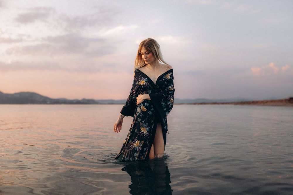 Scenic coastal sunset view with silhouette figure wearing black lace dress standing at ocean's edge during golden hour.