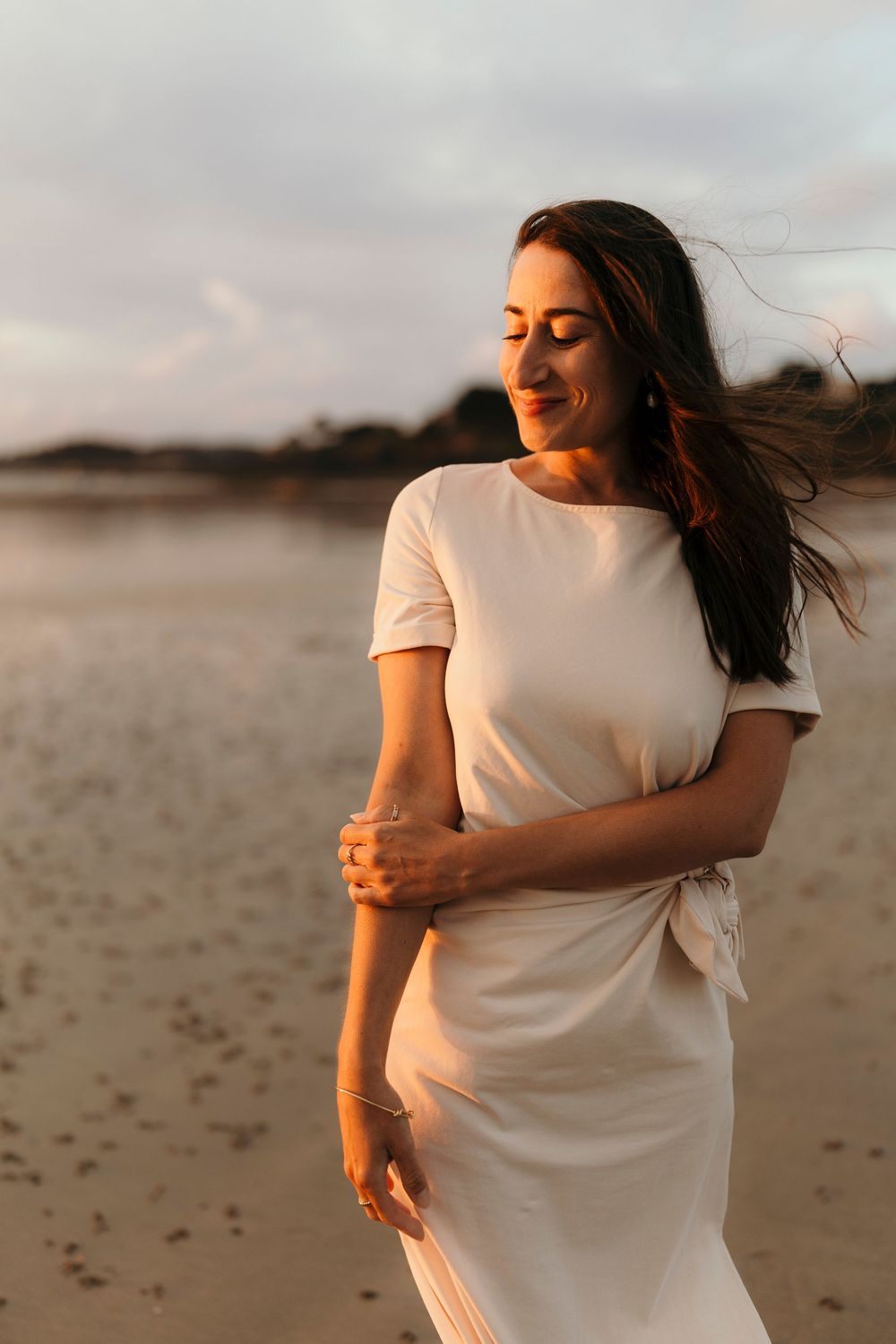 A person in a white dress stands on a beach at sunset with wind blowing through their hair.
