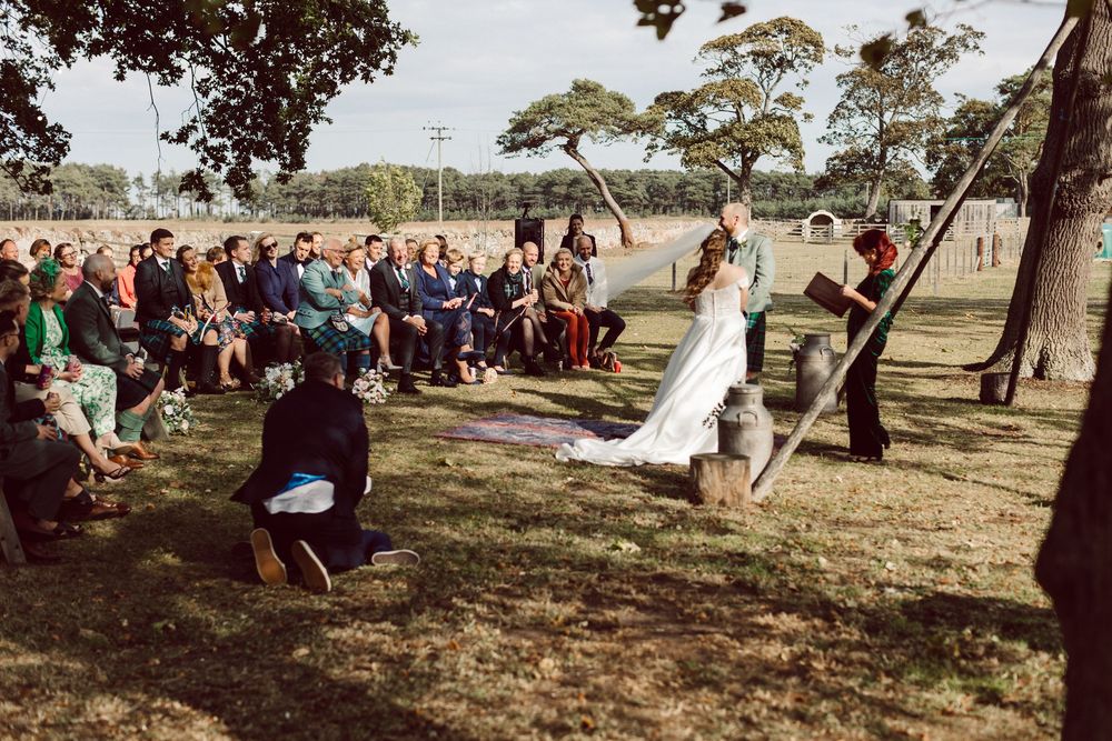 A panoramic view of an outdoor wedding ceremony with guests gathered under trees in a rustic natural setting.