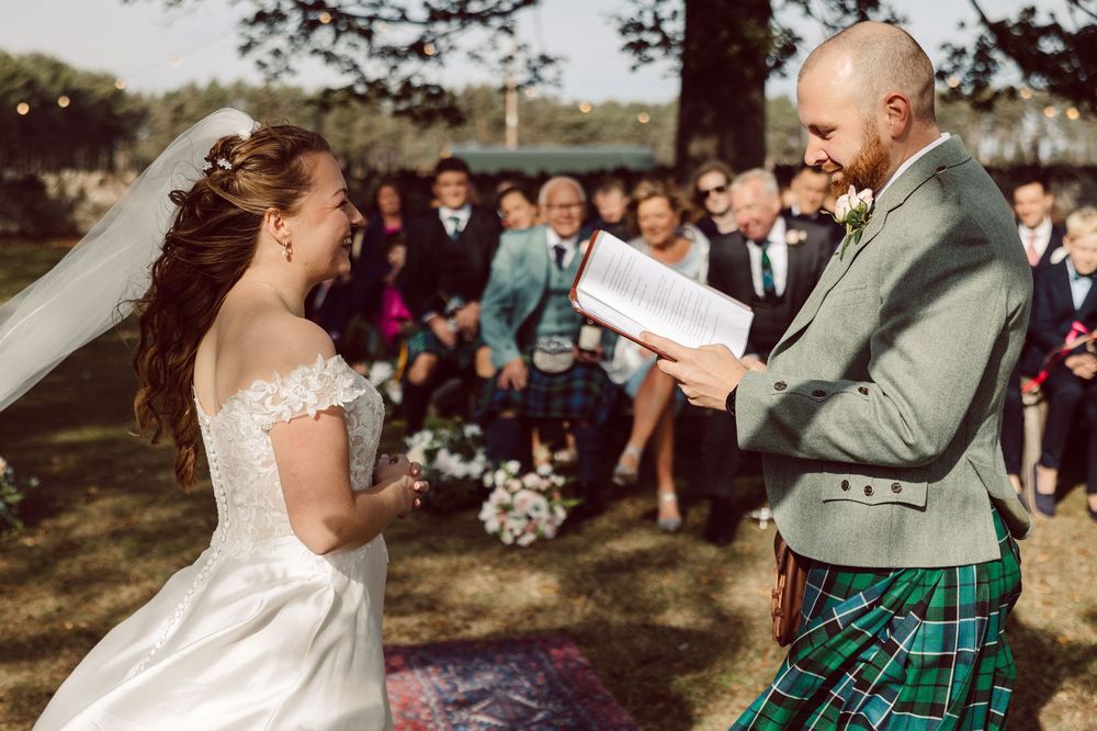 A couple exchanges vows during an outdoor wedding ceremony, with the groom wearing a traditional Scottish kilt.