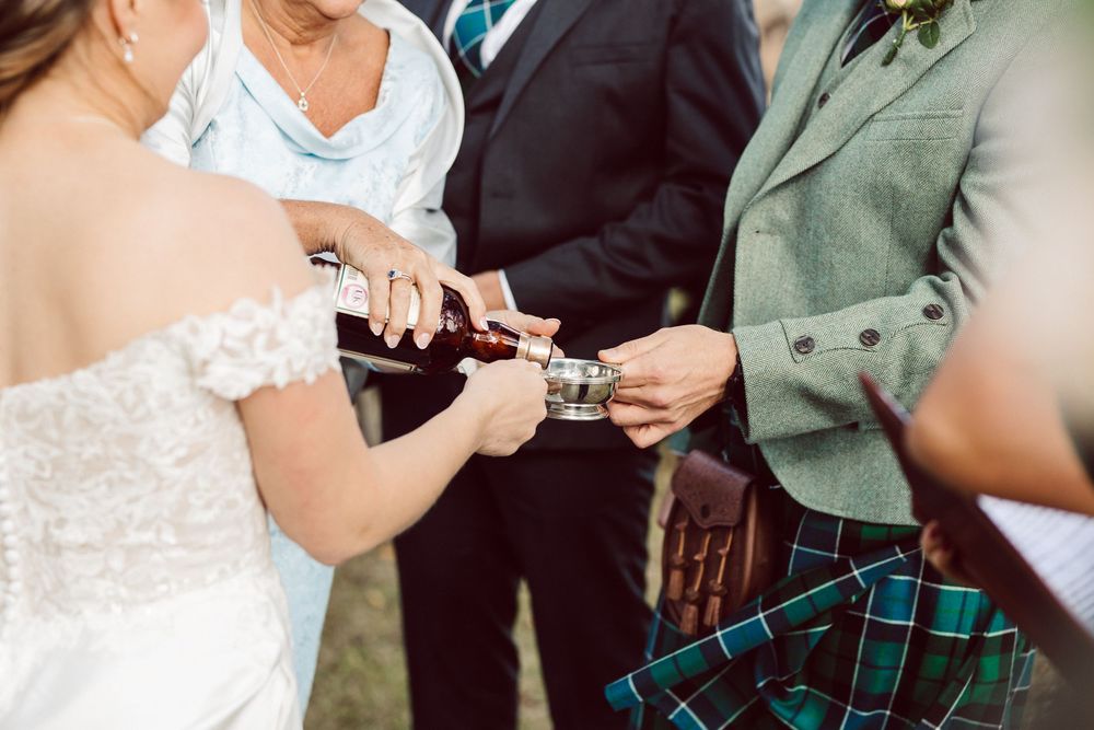 Wedding ceremony participants gather outdoors while exchanging vows and rings in garden setting.