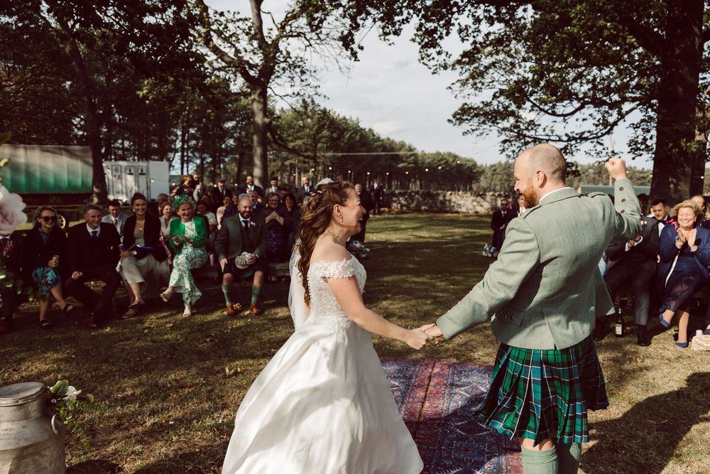 A group of wedding guests dancing together outdoors in a park setting during a sunny celebration.