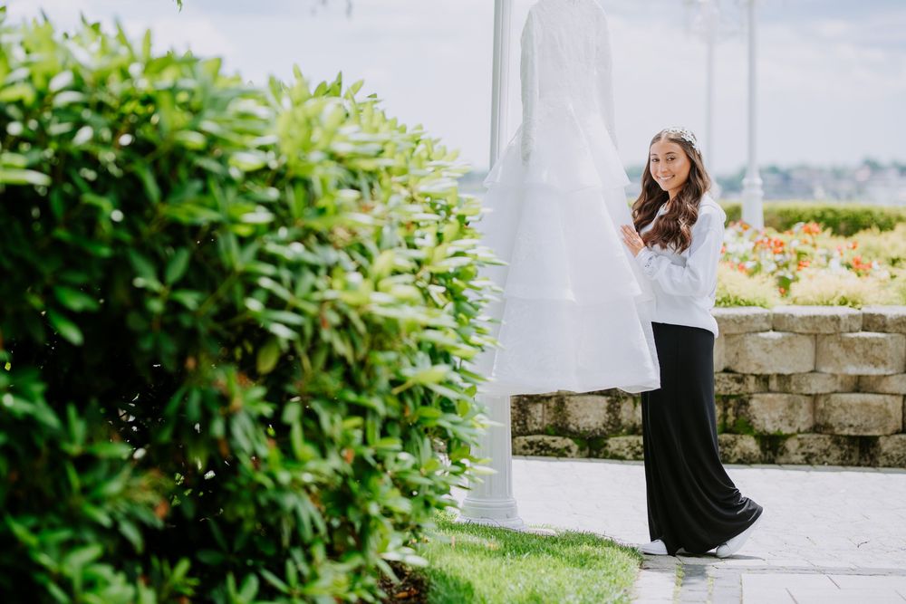 A person in a white top and black pants poses outdoors near lush greenery and a stone wall.