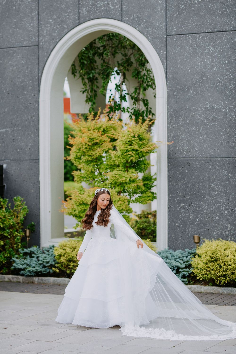 A bride in a flowing white wedding dress poses gracefully against an arched stone wall with lush gardens in the background.