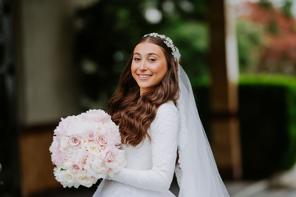 A bride in a white wedding gown and veil holds a pink and white bouquet of flowers in an outdoor garden setting.