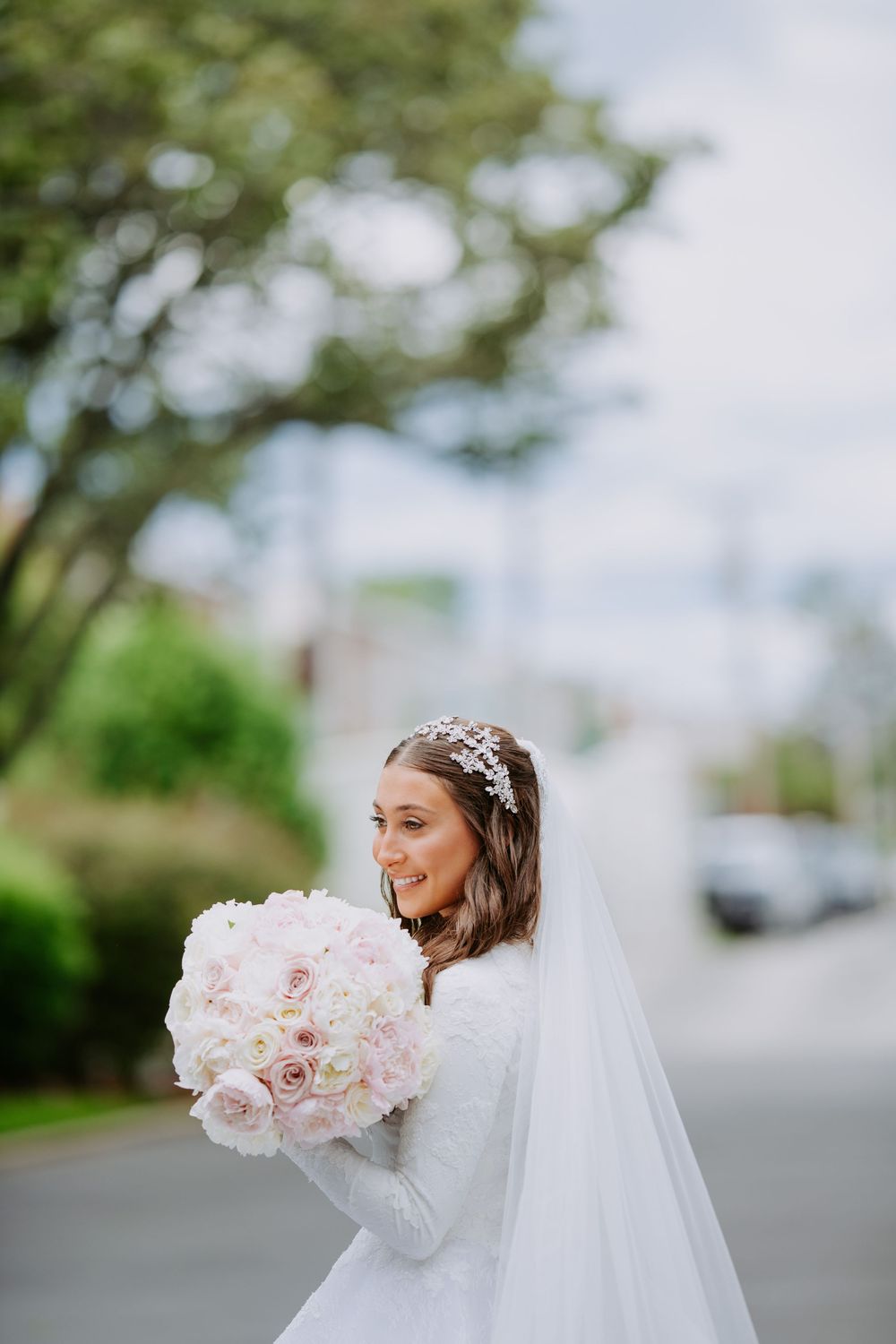 A bride in a white wedding dress and veil holds a beautiful pink rose bouquet on a tree-lined street.