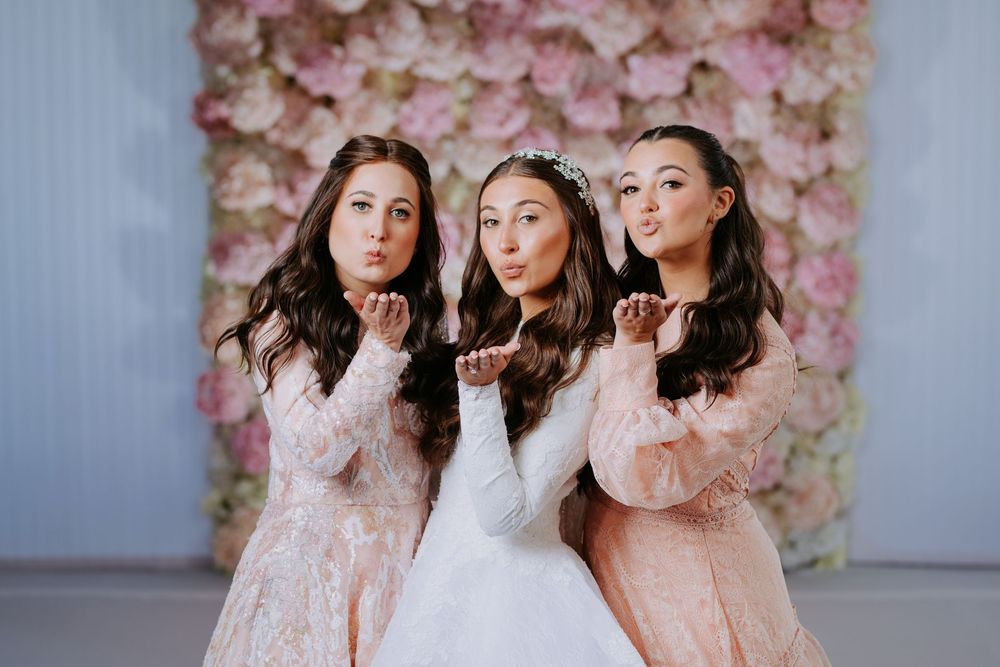 Three women in pink and white lace dresses pose together against a floral backdrop.