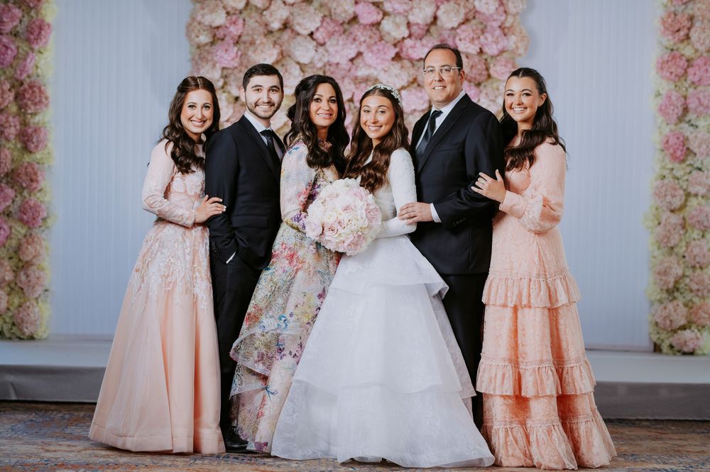 Wedding party poses in front of pink floral wall wearing formal attire in peach and white colors.