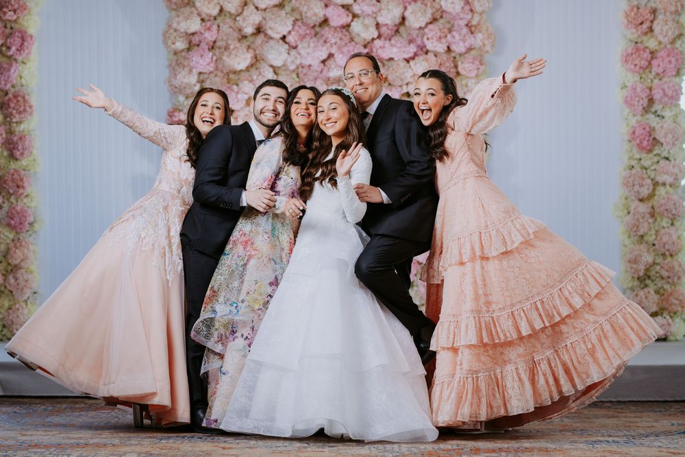 Wedding party in formal attire poses joyfully against floral backdrop wearing pink and black dresses and suits.