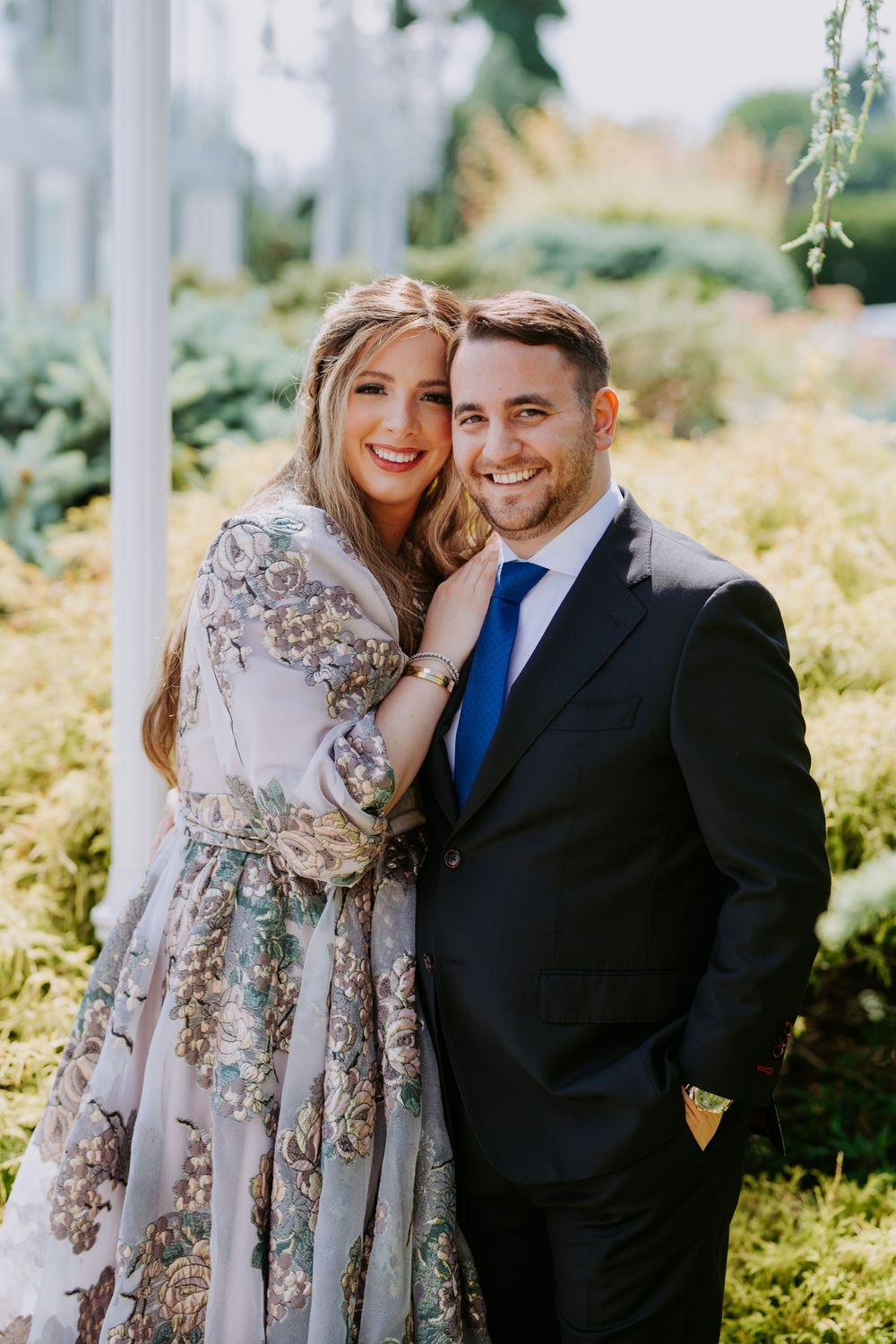 A couple poses together outdoors wearing formal attire with a floral dress and navy suit.