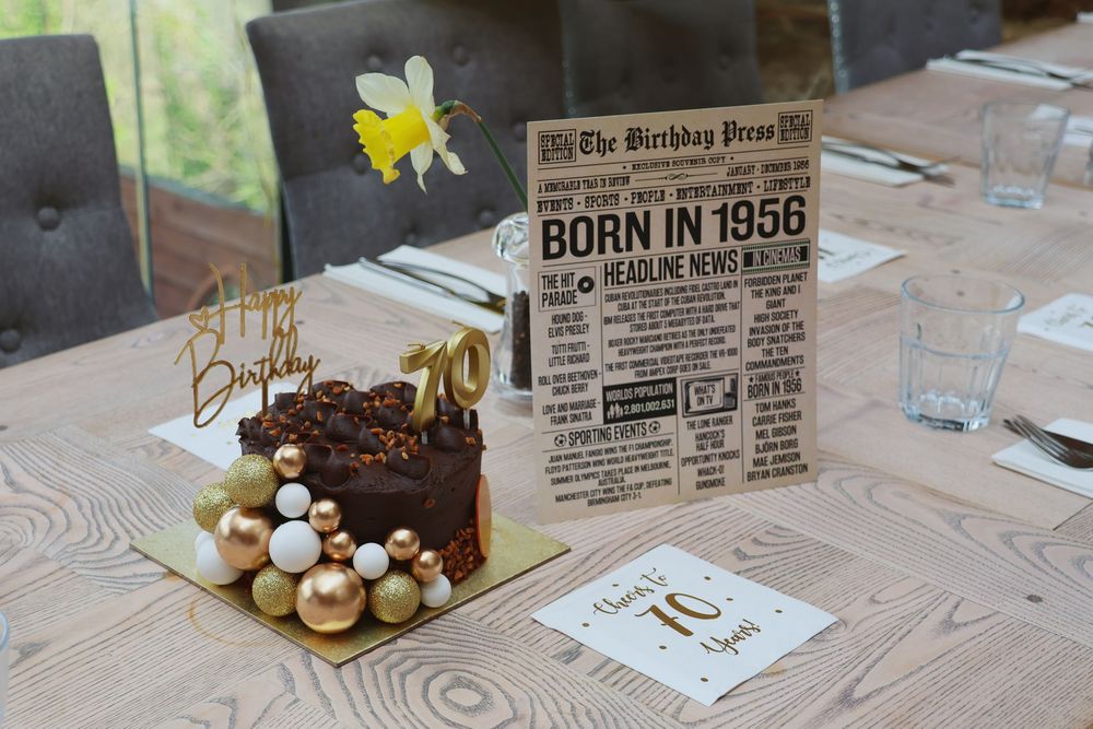 A 70th birthday celebration table with chocolate cake, gold decorations, and a vintage “Born in 1956” newspaper-style card.