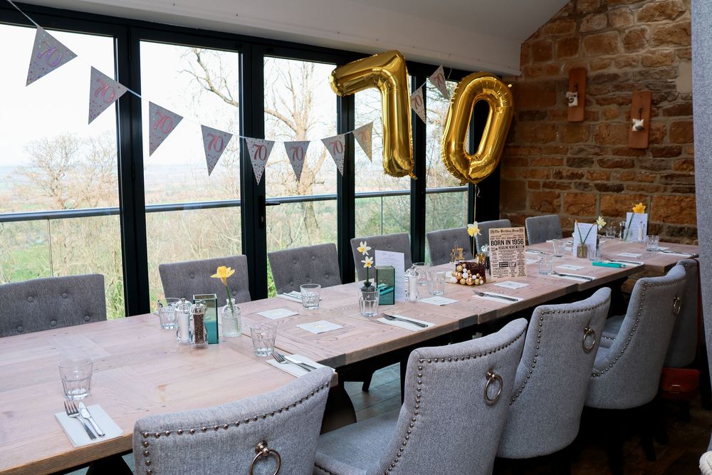 Elegant private dining room with long wooden table, grey chairs, gold 70 balloons, bunting, and scenic window views.